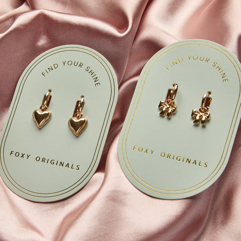 Gold heart-shaped earrings on 'Foxy Originals' cards against a pink satin background