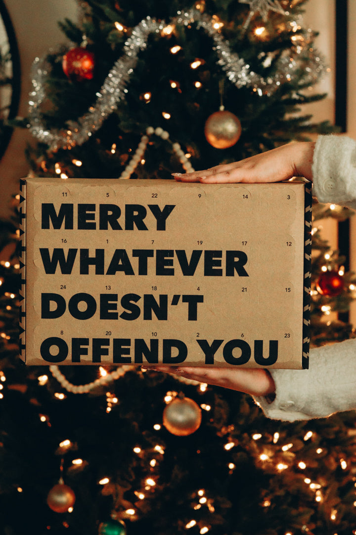 Kraft sign reading Merry Whatever Doesn't Offend You held in front of a lit Christmas tree at The Local Space