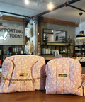 Two pink floral zippered pouches with gold-tone hardware and ribbon ties, displayed on a wooden counter inside a boutique store.