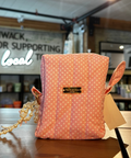 A pink and white polka dot fabric pouch bag with a small gold logo plaque and a pearl-like strap, sitting on a wooden counter inside a store.