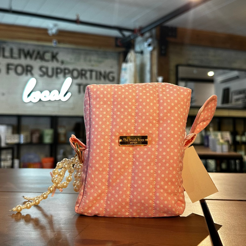 A pink and white polka dot fabric pouch bag with a small gold logo plaque and a pearl-like strap, sitting on a wooden counter inside a store.