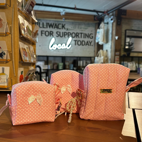 Three pink polka-dot fabric pouches with bow details are displayed on a wooden counter inside a boutique setting.