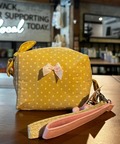 A yellow fabric pouch with white polka dots, a pink bow, and a pink wristlet strap, displayed on a wooden table inside a shop.