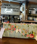 A quilted fabric pencil case with a floral print in sage green, pink, and yellow, resting on a wooden counter inside a shop.