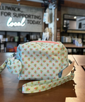 A small fabric pouch with a matching wristlet strap, featuring a light blue and white floral pattern with small pink and yellow accents, resting on a wooden table inside a shop.